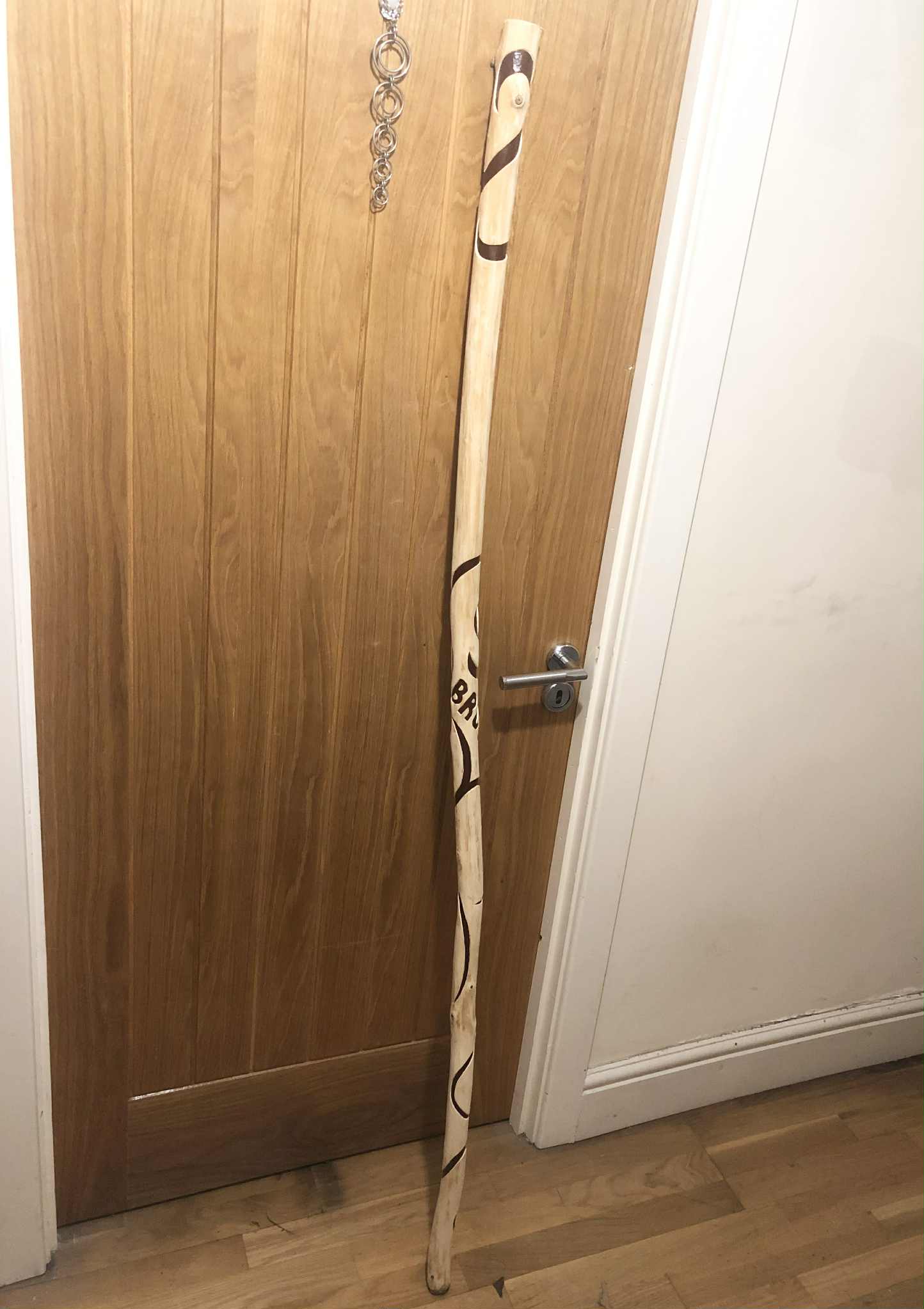 A large, peeled poplar wood staff, with linework carved down the length of it painted in dark reddish brown, leaning against a wooden door.