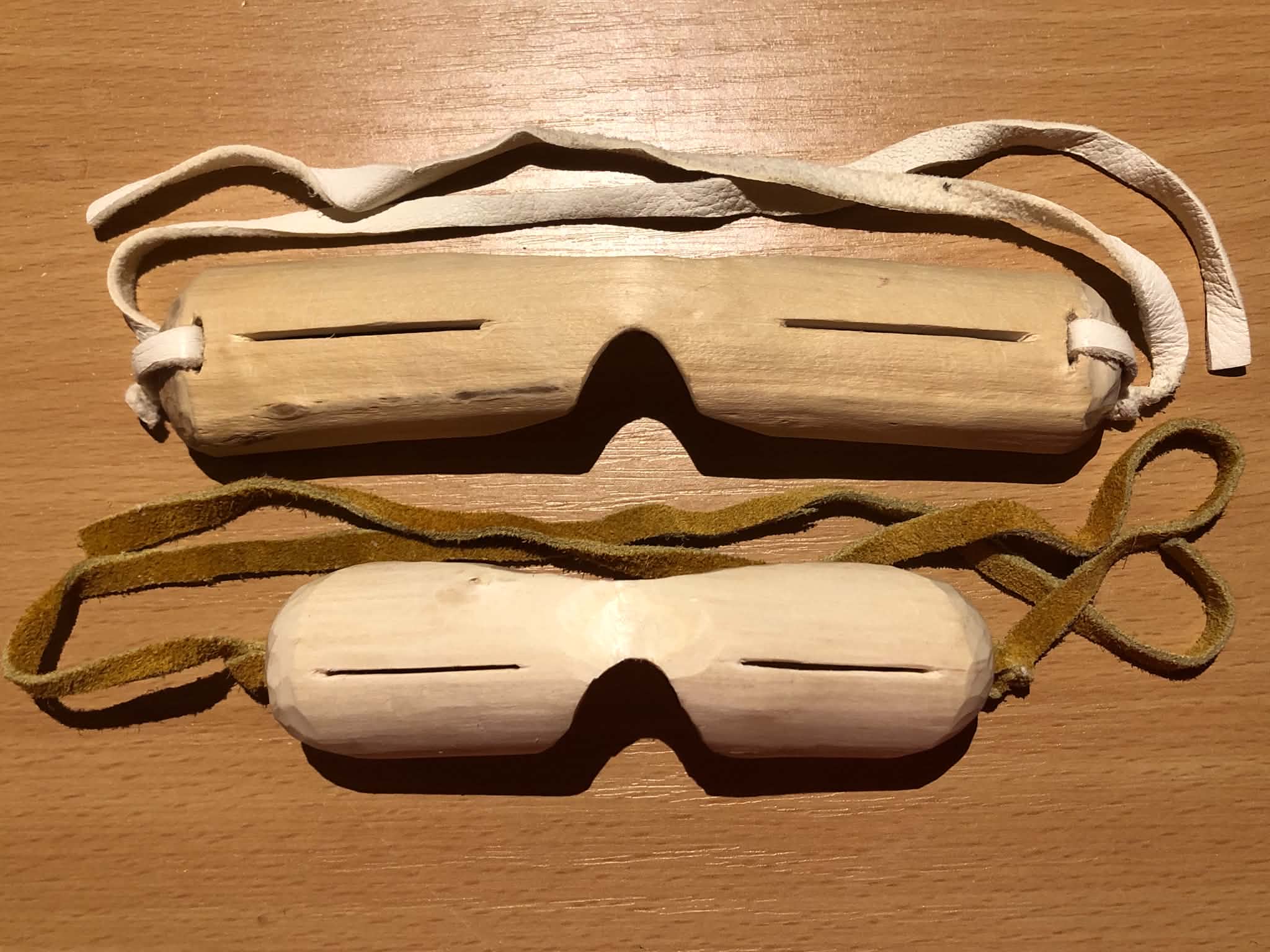 Two pairs of Inuit style snow goggles made from wood, a willow branch with two thin slits cut into it to see through, a nose and forehead groove, and leather straps to tie them around your head.