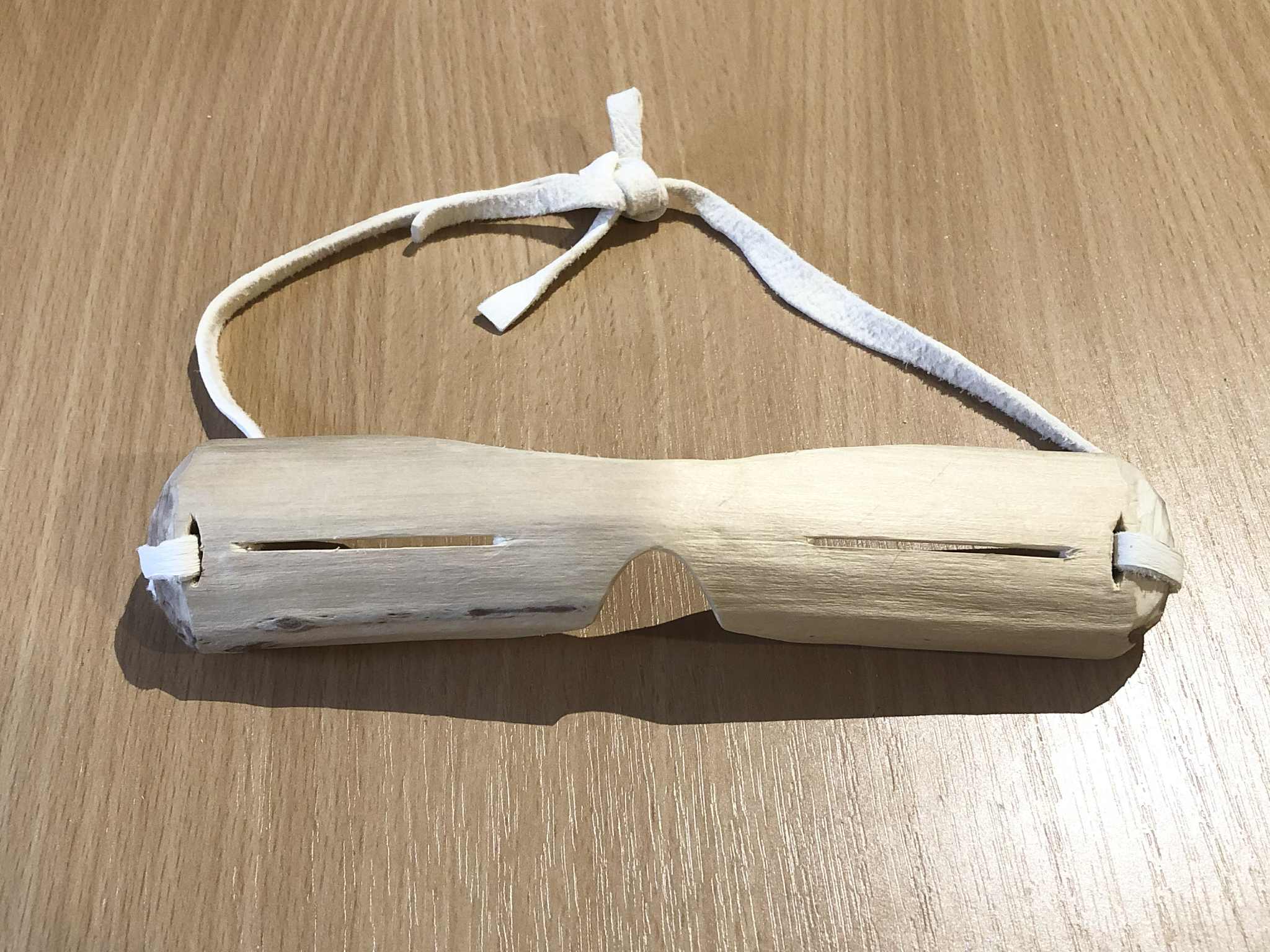 A pair of Inuit style snow goggles made from wood, a white willow branch with two thin slits cut into it to see through, a nose and forehead groove, and white leather straps to tie it around your head.