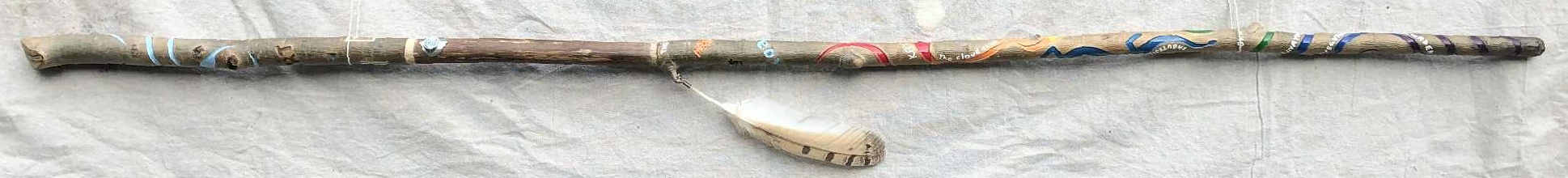 A colourful staff positioned in front of a canvas backdrop. Curved lines of different colours, light blue at the top, and a rainbow running down through the body, with different words and designs carved amongst the curves, such as meow, koa, Oka, and others, as well as a large blue dragon, and a metal staff marker. A large light brown feather hangs from an accessory stick on the side below the grip.