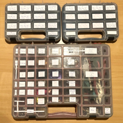 Three craft organizer cases, two smaller black ones and a larger red one beneath, with neat labels over the majority of the slots.