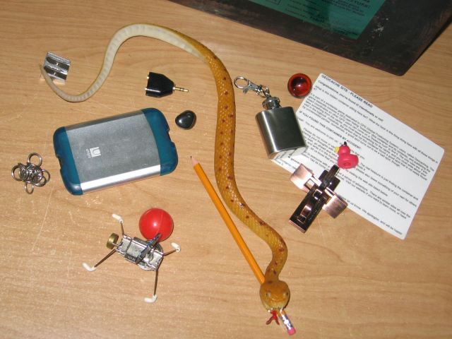 The tradeables inside of the geocache, a chainmaille ball, a windup toy, a plastic card holder, a rubber snake, a tiny little flask, a metal puzzle, a marble, and a few more odds and ends, and the geocache site instruction sheet.