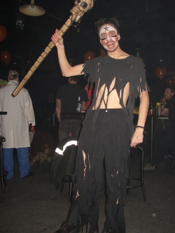 Kabutroid at a halloween event, wearing shredded pants and a shirt, with facepaint and her shaman stick.