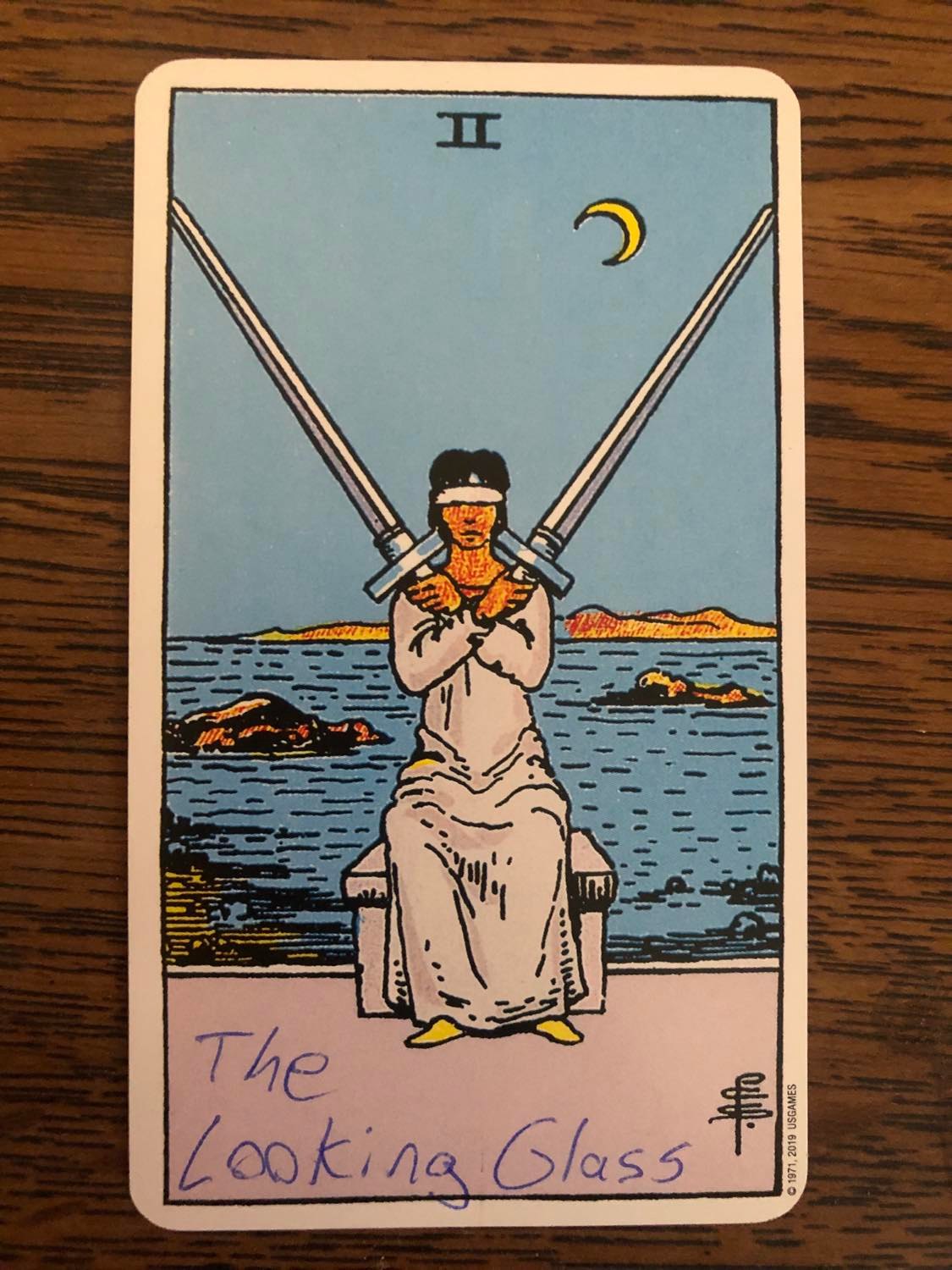 The two of swords card with the looking glass written on it, laying on a wooden desk.