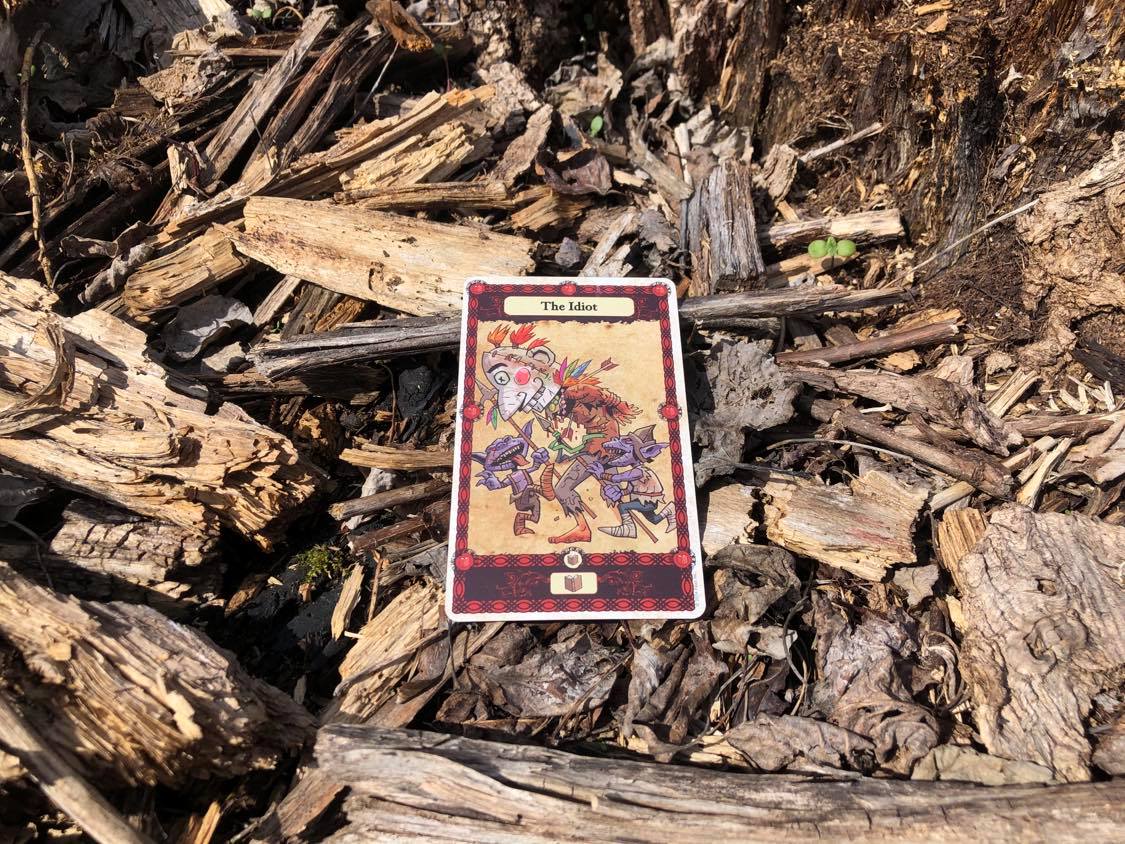 A person wearing a jester mask surrounded by goblins, the card laying on the forest debris.