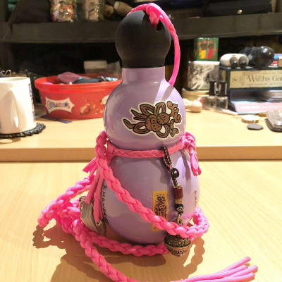 A light pink purple lavender coloured, gourd shaped bottle, wrapped in pink cording, with a tiny pen, notepad, and an ornate bell hanging from the side.