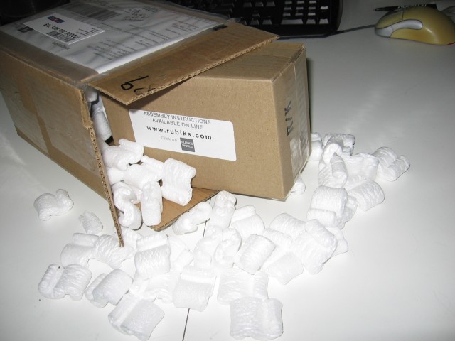 The cardboard box opened on one side, packing peanuts and a smaller box inside spilling out, a computer mouse and keyboard in the background.