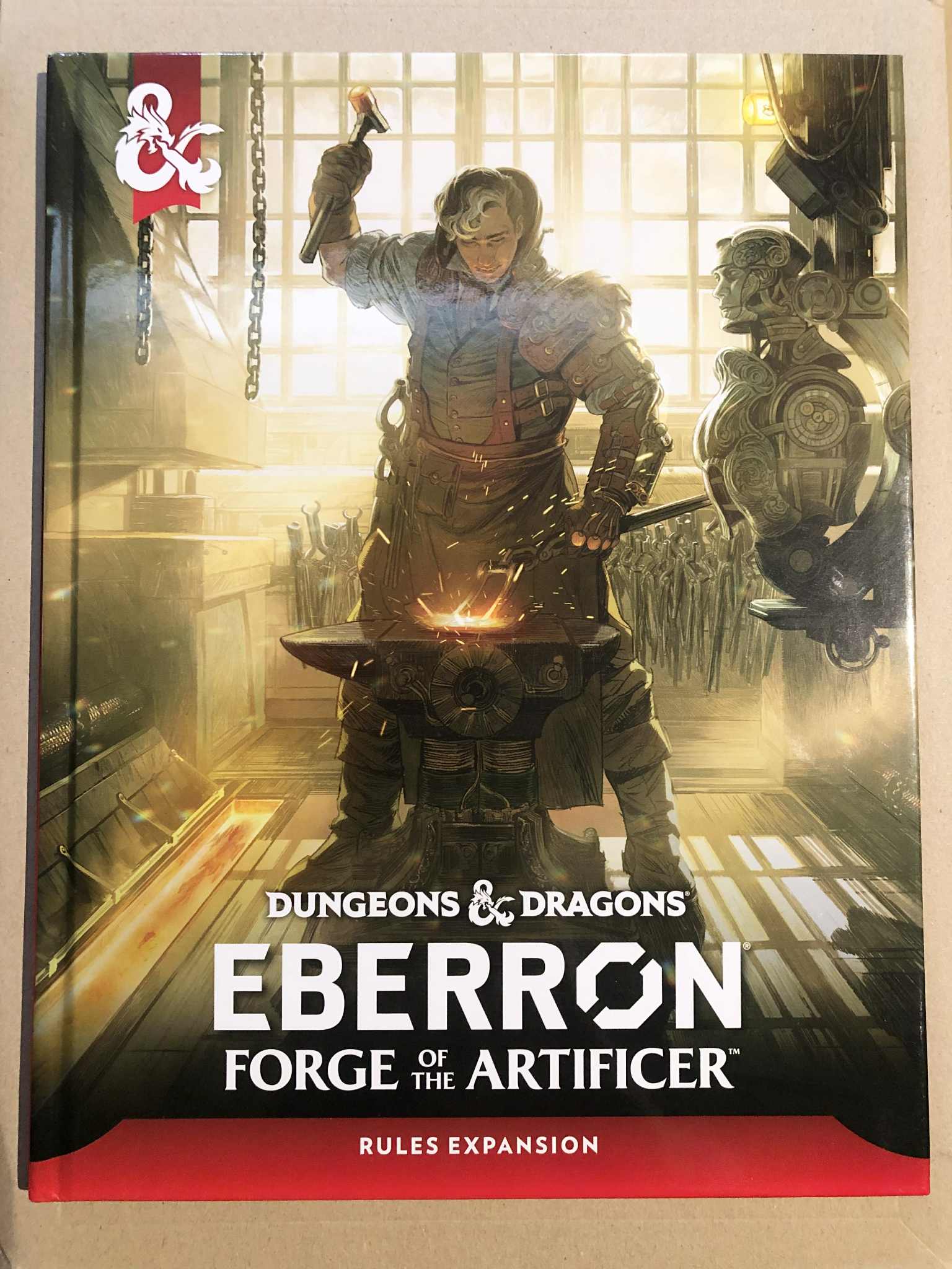 The d and d book Eberron, forge of the artificer, a hardcover book with a cover featuring a man working at a modified anvil, with much wiring hooked to it, a half built robot beside him. Behind are dozens of clamps and other blacksmithing tools, and the sunlight shines in from a window behind him.