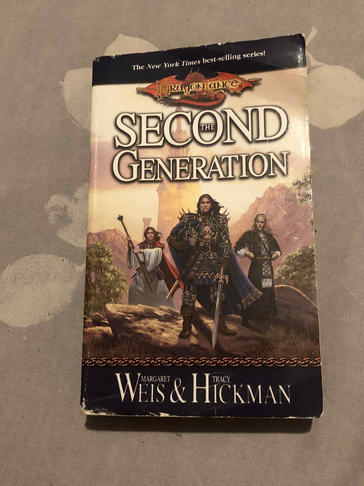 A book titled Dragonlance The Second Generation, featuring a white robe wearing human cleric holding a staff, an elf wearing armour and standing with their hands on their hips, and a human wearing black spiked armour holding a skull helmet in one hand and an ornate sword in the other.
