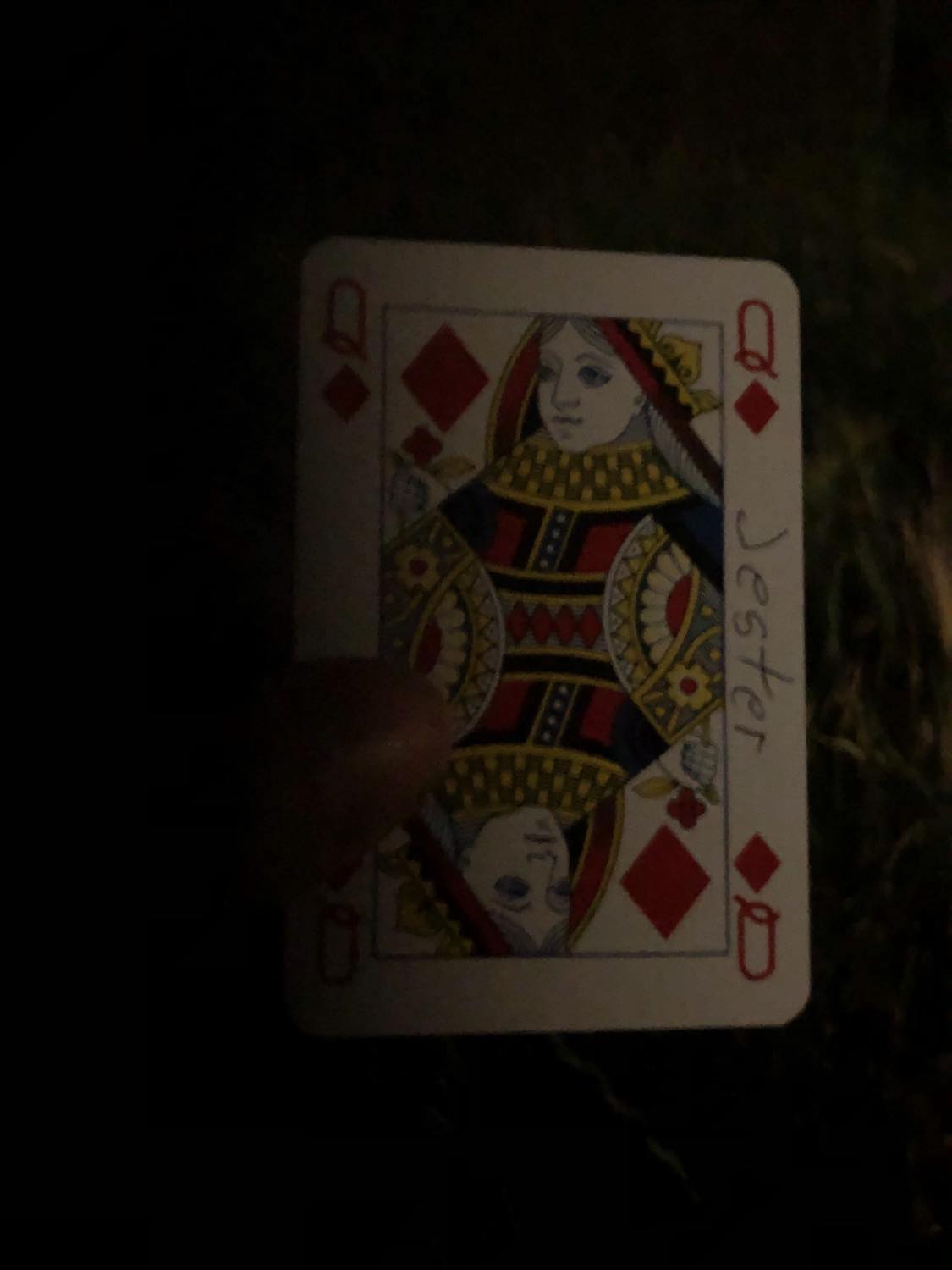The queen of diamonds with the word Jester written on it, held in the barely lit dark.